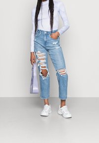 Jean déchirés bleu clair avec de multiples détails de vieillissement, associés à un haut blanc à manches longues et des baskets blanches. Tenant un sac lavande.