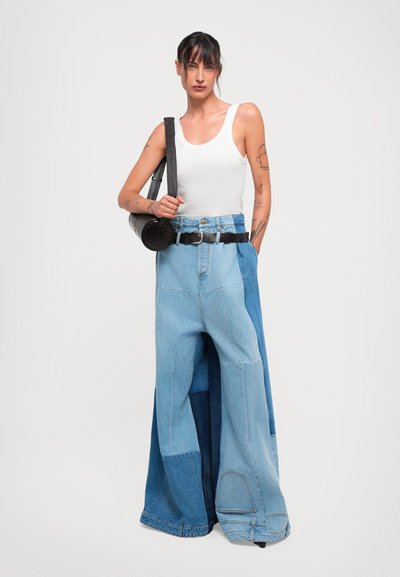 Kobieta z tatuażami ubrana w biały top na ramiączkach, szerokie jeansy w stylu patchwork, czarny pasek i trzymająca czarną torbę na ramię na tle białym.