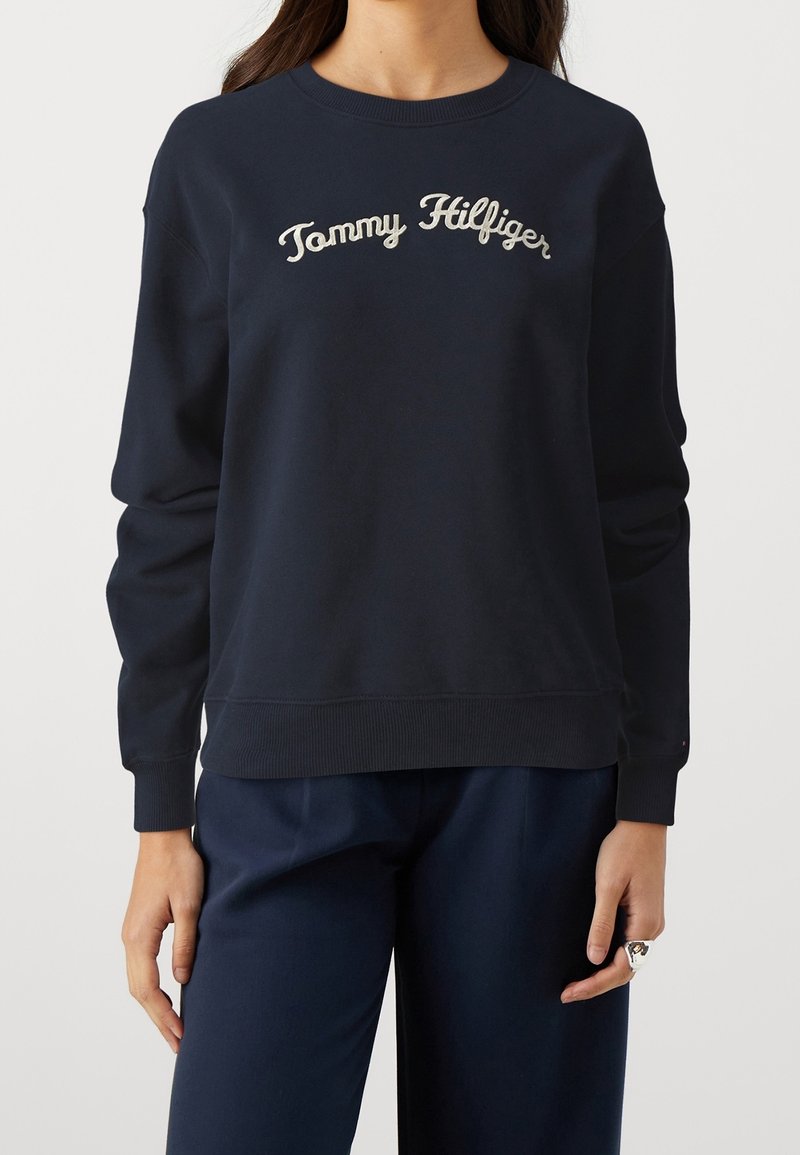 Femme portant un sweat-shirt bleu marine avec « Tommy Hilfiger » brodé en lettres cursives blanches sur la poitrine, associé à un pantalon bleu marine assorti.