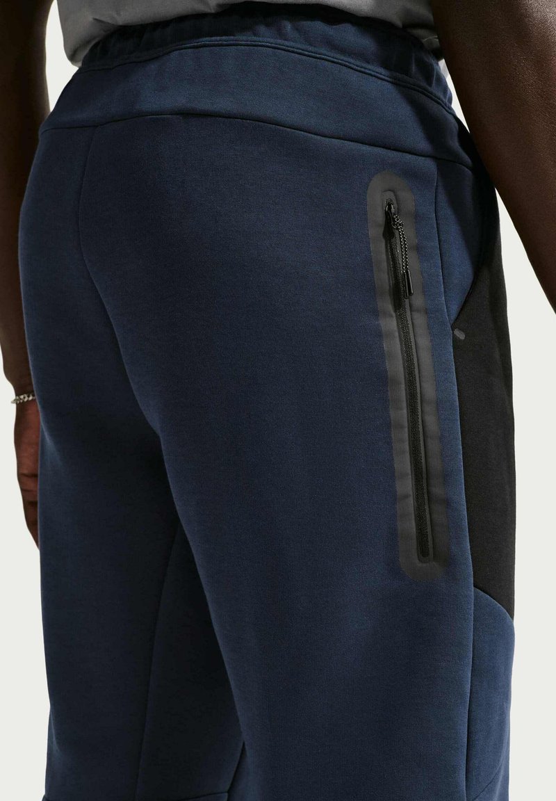 Pantalones deportivos azul marino con un panel lateral negro texturizado, que incluye un bolsillo con cremallera en la pierna derecha. Cintura elástica para mayor comodidad.