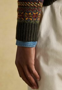 Sötétzöld, kötött pulóverujj, több színű geometrikus mintákkal. Világoskék farmering felett viselve, kéz fehér szöveten pihentetve.