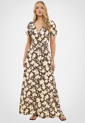 Frau trägt ein braunes Wickelkleid mit cremefarbenem Blumenmuster, Puffärmeln und Sandalen, steht vor einem schlichten hellen Hintergrund.