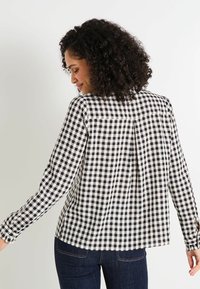 Blusa de manga larga en patrón de cuadros en blanco y negro, con pliegue en la parte posterior, tejido texturizado y detalles de puños abotonados.