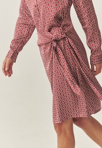 Rochie roșie cu model geometric, mâneci lungi și detaliu cu nod în talie. Material neted cu un luciu subtil și croială de lungime medie.