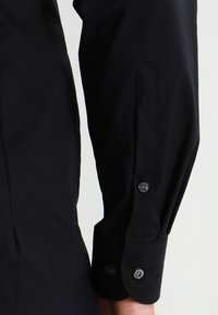 La chemise noire à manches longues présente une coupe ajustée, une texture lisse, un poignet avec deux boutons et un détail d'encoche subtil près du poignet.