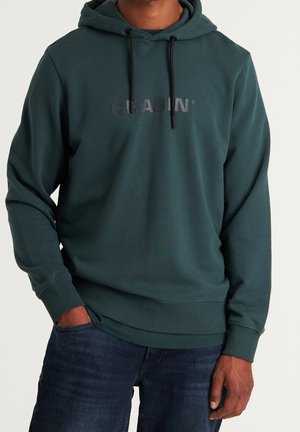 Personne portant un sweat à capuche vert foncé avec le texte noir "CHASIN'" et des cordons noirs, associé à un jean bleu foncé, mains détendues le long du corps.