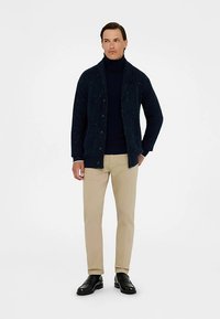 Marineblauw vest met knopen en een textuurrijk gebreid ontwerp, gedragen over een donkere coltrui, gecombineerd met beige broek en zwarte nette schoenen.