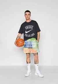 Sort T-shirt med hvid tekst og Nike-logo, flerfarvede mesh-shorts, hvide sko, mens der holdes en basketball. Accentueret med tatoveringer og pandebånd.
