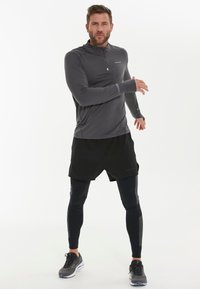 Grijze lange mouw sporttop met halve rits, zwarte shorts, zwarte compressieleggingen en zwarte hardloopschoenen. Gevoelige stofdetails zichtbaar.