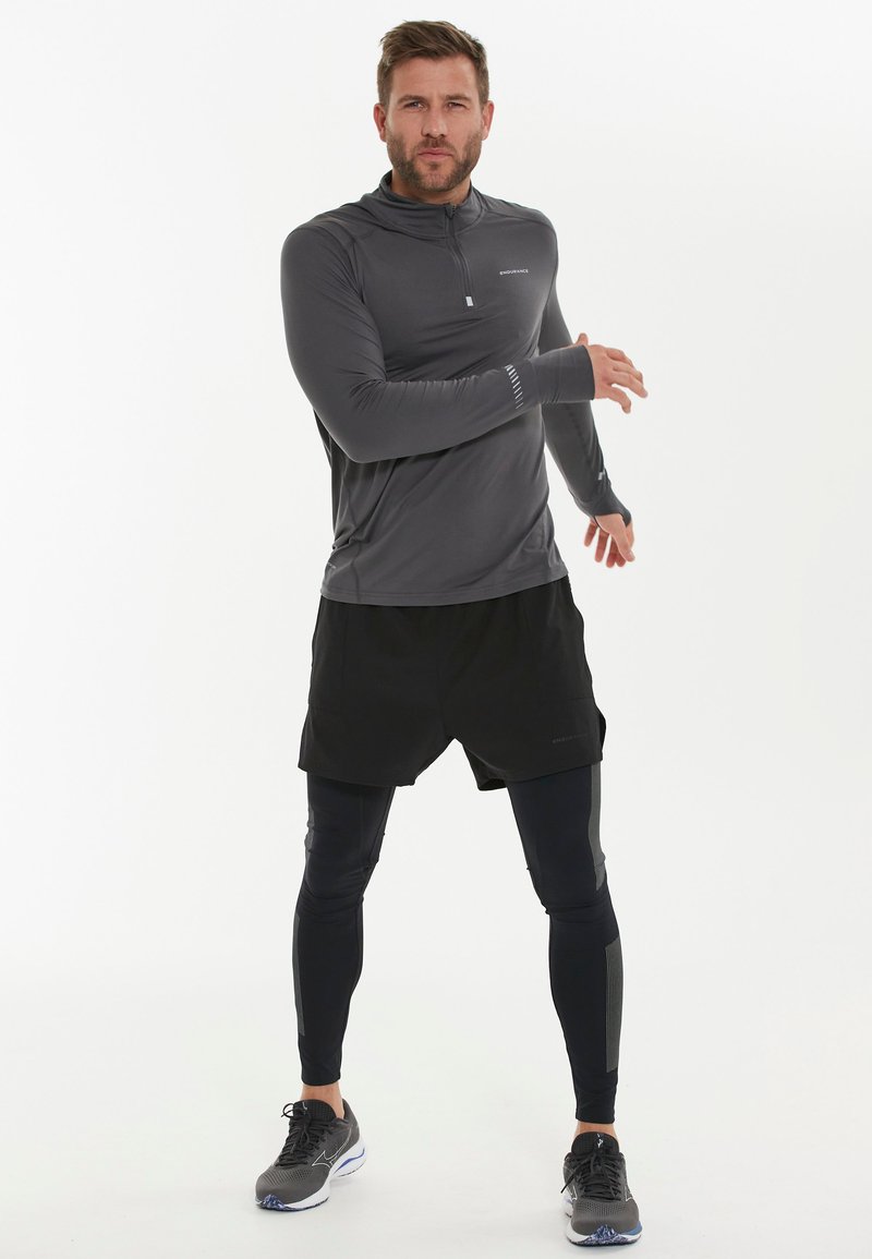 Grijze lange mouw sporttop met halve rits, zwarte shorts, zwarte compressieleggingen en zwarte hardloopschoenen. Gevoelige stofdetails zichtbaar.