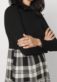 Anillo de oro con una piedra verde en una mano, vistiendo una blusa negra con textura de canalé, con los brazos cruzados sobre una falda a cuadros en blanco y negro.