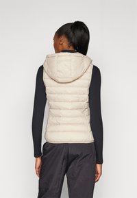 Weste aus leichtem Beige mit Kapuze, ausgestattet mit einem gesteppten Muster. Ärmelloses Design, weiche Textur und eine figurnahe Silhouette.