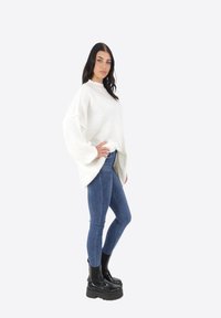 Maglione bianco a maglia grossa con spalle basse, abbinato a jeans blu aderenti e stivali neri con plateau, caratterizzato da una texture liscia e una vestibilità rilassata.