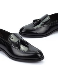 Mocasines de cuero negro con un acabado brillante, borlas decorativas en la parte frontal y costuras a lo largo del borde. Suela de goma.