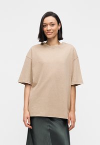 T-shirt beige oversize à manches courtes et col rond côtelé ; porté avec une jupe en satin foncé, texture lisse, design minimaliste.