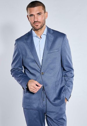 Mann mit kurzen Haaren und Bart, der einen blauen Anzug und ein hellblaues Hemd trägt, steht mit einer Hand in der Tasche und einer Hand in der Nähe der Jacke.