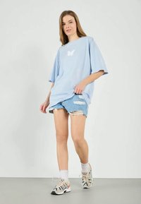 Světle modré oversized tričko s bílou grafikou motýla, kombinované s distressed denimovými šortkami a sportovními teniskami.