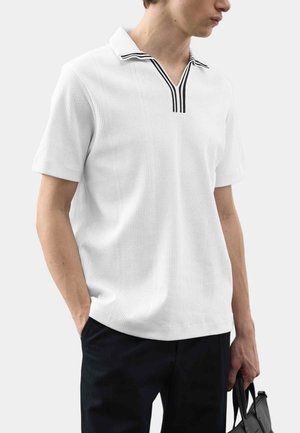 Mężczyzna ubrany w biały, krótkorękawowy sweter polo z kołnierzykiem w czarne paski, czarne spodnie, trzymający czarną torbę w dłoni.