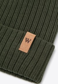 Dunkelgrüne, gerippte Strickmütze mit umgeschlagenem Bündchen und einem braunen Lederetikett mit einem stilisierten "W". Die Textur wirkt weich und warm.