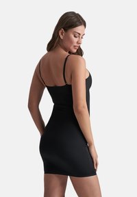 Una donna di spalle che indossa un vestito attillato nero senza maniche con spalline sottili, su uno sfondo chiaro e semplice.
