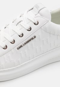 Vit sneaker med texturerad syntetisk ovandel, platta vita snören, silverdetaljer och präglad logotypdetalj på sidan.