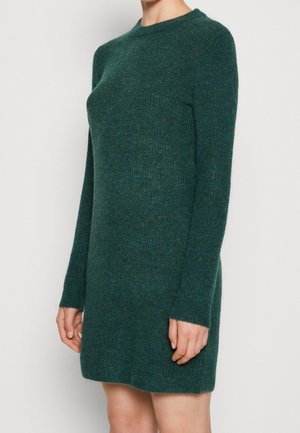 Robe tricotée vert foncé à manches longues et encolure ronde. La texture est douce et légèrement rugueuse, avec une silhouette ajustée.
