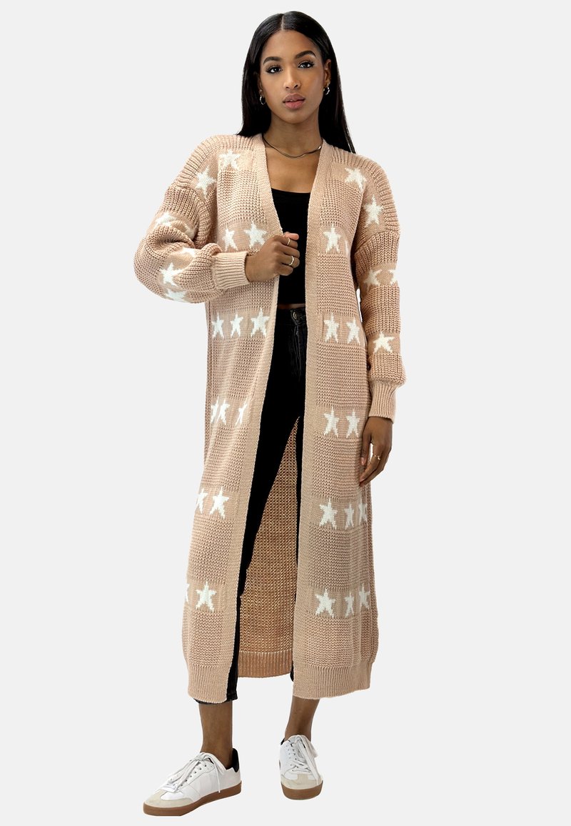 Cárdigan largo de punto en color beige claro con patrones de estrellas blancas, de apertura frontal, con puños acanalados y tejido texturizado. Llevado sobre una blusa negra y vaqueros.