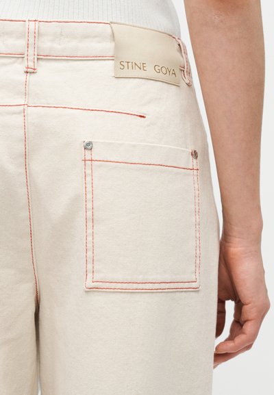 Beigen väriset denimin housut, joissa on punaiset ompeleet ja suorakulmainen takatasku, jossa on vyötäröllä kohokuvioitu "STINE GOYA" -merkki.