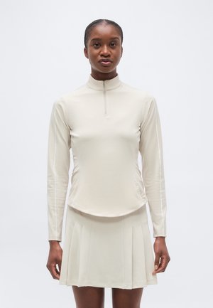 Γυναίκα που φορά ένα μακρυμάνικο μπεζ τοπ με φερμουάρ και ταιριαστή πλισέ φούστα, στέκεται μπροστά σε ένα απλό φόντο.