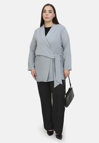 Top avvolgente grigio chiaro con maniche lunghe, tessuto testurizzato e dettaglio in vita annodato, abbinato a pantaloni neri a gamba larga e a una borsa nera.