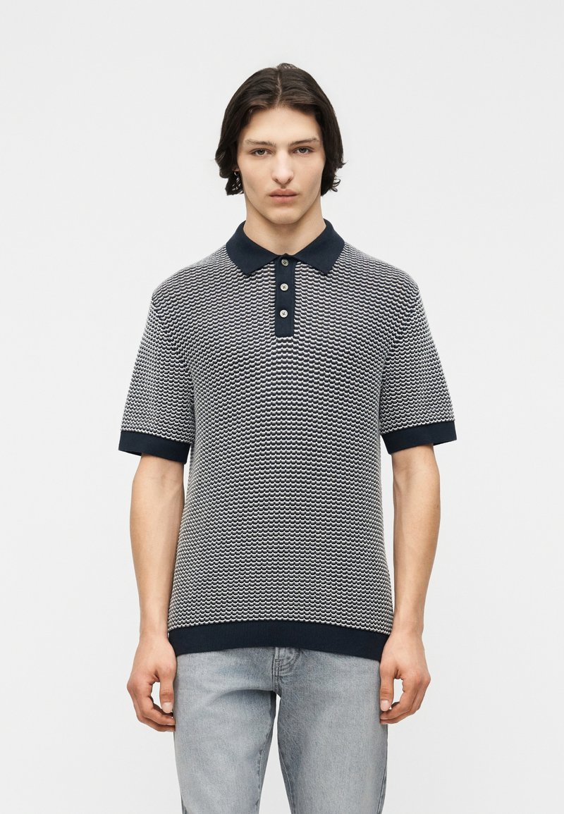 Jeune homme aux cheveux mi-longs et foncés, portant un polo à manches courtes bleu marine et blanc à motifs, et un jean bleu clair.