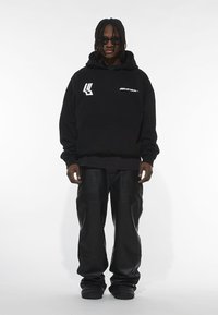 Sort bomulds hoodie med forlommer og hvidt logo. Parret med løse sorte læderbukser. Modellen bærer solbriller. Minimalistisk design.