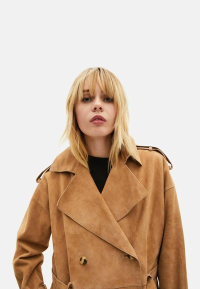 Gabardina de ante color camello con frente de doble botonadura, solapas anchas y charreteras en los hombros. Textura suave y diseño estructurado.
