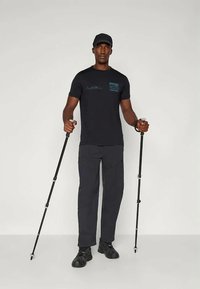 Čierne tričko s grafickým motívom hory, tmavé cargo nohavice a čierne turistické palice v stoji na neutrálnom pozadí.