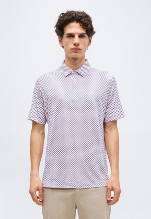 Jeune homme aux cheveux bouclés portant un polo blanc à manches courtes avec un petit motif de cœurs sombres et un pantalon beige, debout devant un fond uni.