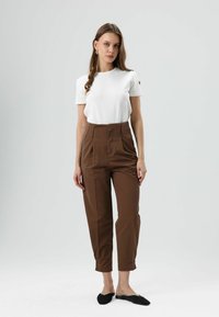 Wit T-shirt gecombineerd met hoge taille bruine broek. De broek heeft een gladde textuur en subtiele plooien, aangevuld met zwarte instapschoenen.