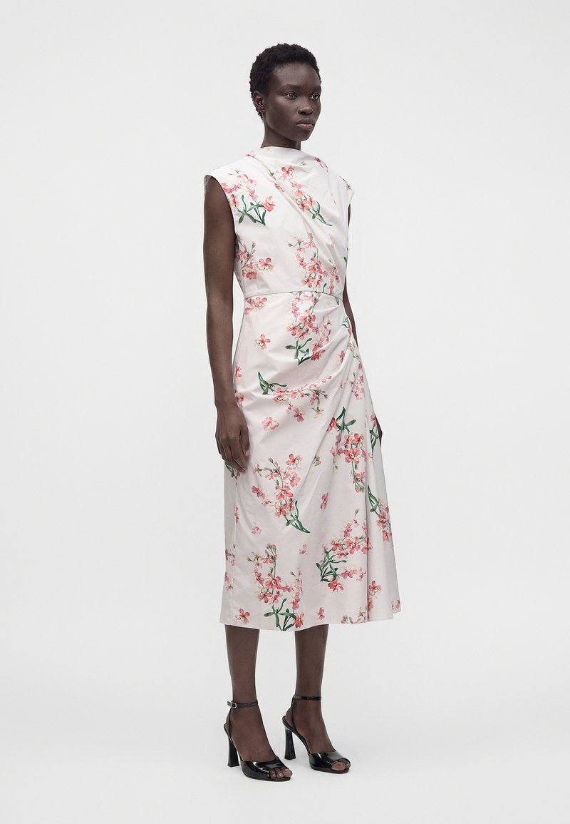 Woman standing wearing a sleeveless white midi dress with pink floral print and black open-toe high heels against a plain light background.