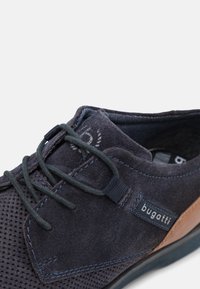 Chaussures en daim marine avec détails en mesh, lacets plats, accents marron contrastants et piqure bleue. Présente une étiquette de marque sur le côté.