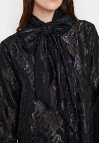Svart blus med en strukturerad, transparent tyg med ett mönstrat mönster. Inkluderar en knytbanddetalj vid halsen som är gjord av samma material.