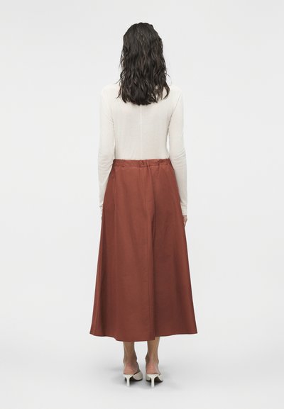 Γυναίκα με σγουρά σκούρα μαλλιά που φοράει λευκό τοπ με μακριά μανίκια, φούστα midi σκουριασμένου χρώματος και άσπρα παπούτσια με τακούνι, στέκεται γυρισμένη προς τα πίσω.