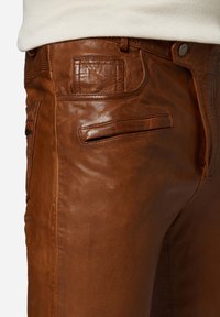 Bruine leren broek met een gladde textuur, voorzien van een rits aan de voorkant, een metalen knoop en meerdere zakken, inclusief details met kleppen aan de achterkant.
