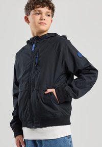 Musta kevyt takki hupulla, vetoketjulla edessä, kahdella sivutaskulla ja sinisillä yksityiskohdilla. Varustettu elastisilla hihansuilla ja rento istuvuus.