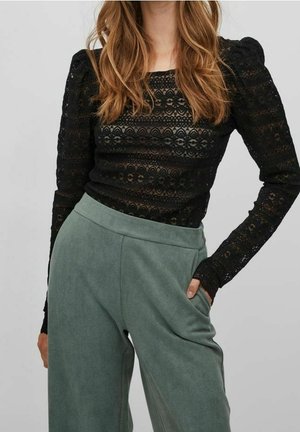 Femme portant un haut en dentelle noir à manches longues avec des épaules bouffantes et un pantalon vert ample taille haute, main dans la poche sur un fond uni.