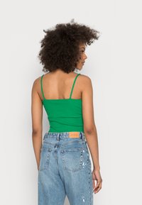 Camisola verde com nervuras e alças finas, combinada com jeans azul desbotado de cintura alta, apresentando uma etiqueta bege na cintura.