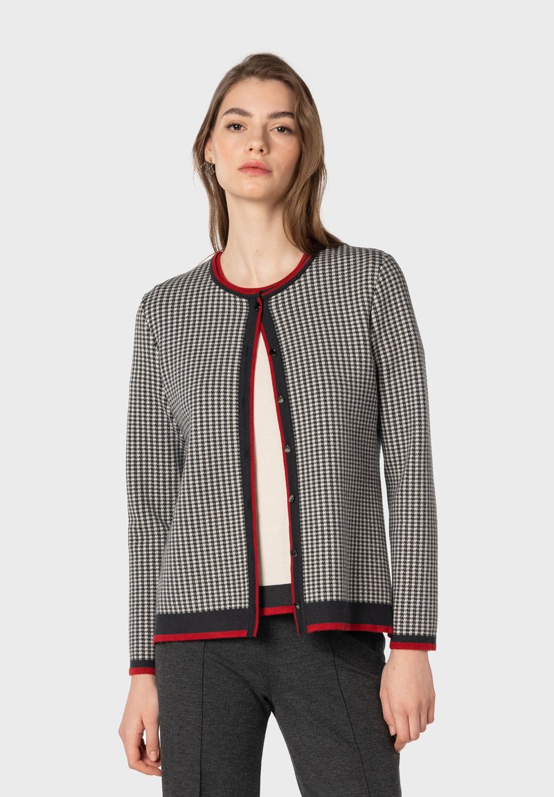 Cardigan lavorato a maglia in pattern houndstooth nero e crema, con bordi ribattuti rossi e bottoni neri. Presenta un collo rotondo e maniche lunghe.