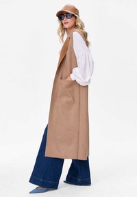 Beżowy bezrękawnik z kieszeniami, zestawiony z białą bluzką, szerokimi dżinsowymi spodniami i brązową czapką. Charakteryzuje się gładką fakturą i prostymi liniami.
