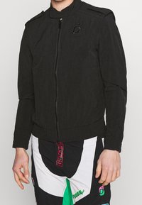 Černá bomber bunda se zapínáním na zip, vysokým límcem a ramenními epauletami. Má strukturovaný vzhled a vyšívané logo na hrudi.