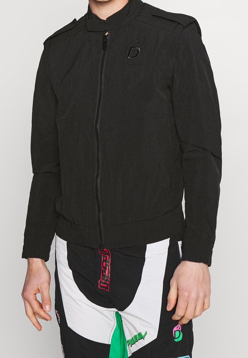 Černá bomber bunda se zapínáním na zip, vysokým límcem a ramenními epauletami. Má strukturovaný vzhled a vyšívané logo na hrudi.