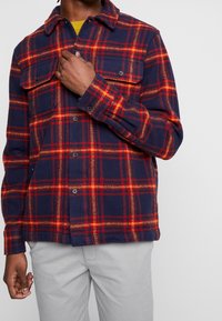 Camicia in flanella a quadri blu navy e rossa con abbottonatura frontale, due tasche sul petto e colletto. Il tessuto ha un aspetto morbido e testurizzato.