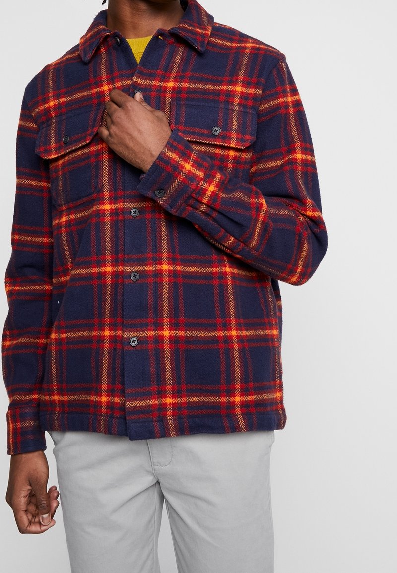 Camicia in flanella a quadri blu navy e rossa con abbottonatura frontale, due tasche sul petto e colletto. Il tessuto ha un aspetto morbido e testurizzato.